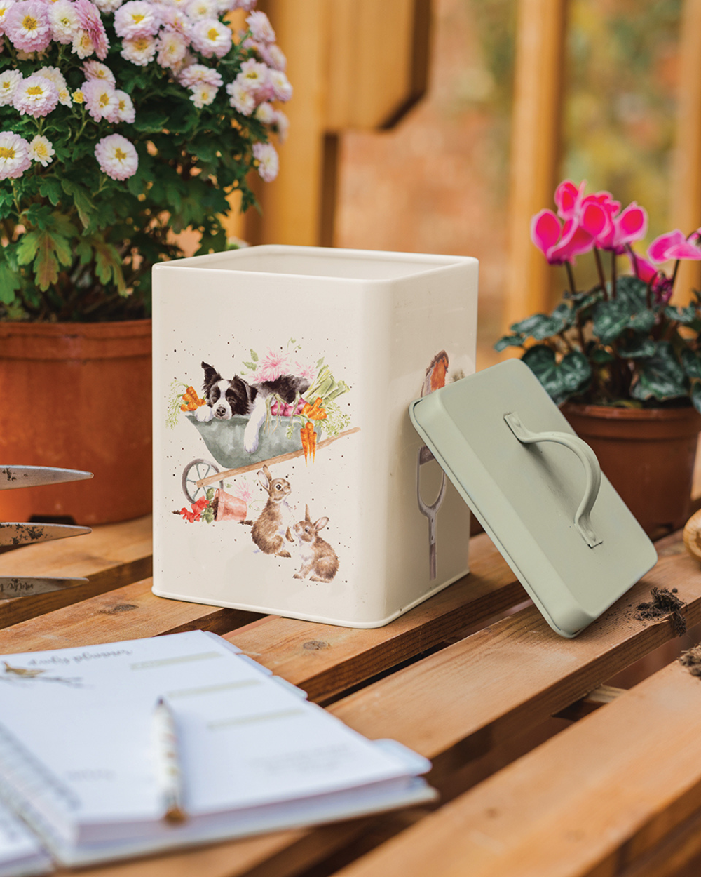 Dekorierte Garten Vorratsdose von Wrendale Designs auf Holzbank zwischen Blumentöpfen, geöffnet mit grünem Deckel