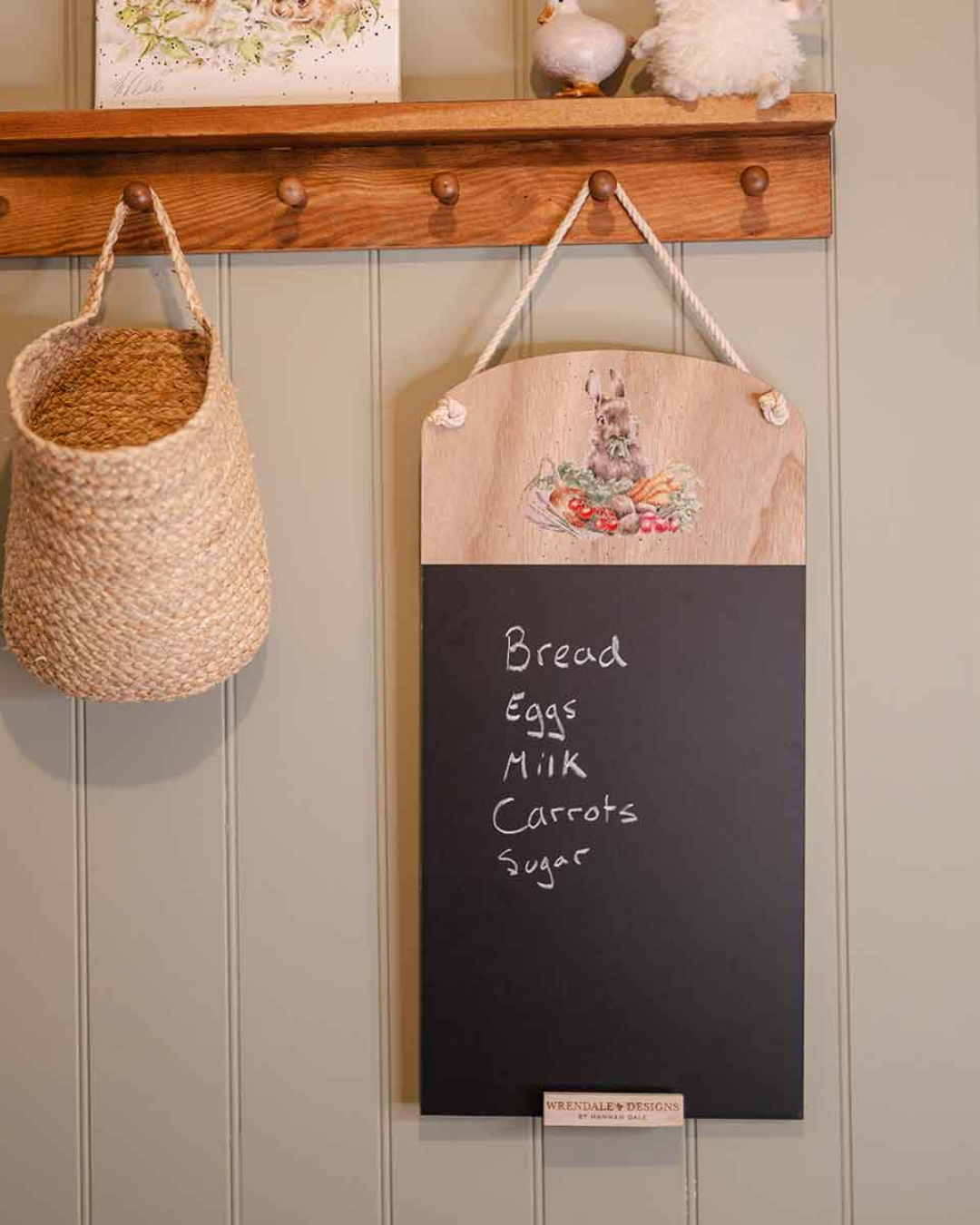 An der Wand hängende Kreidetafel mit Gemüsehase und handgeschriebener Einkaufsliste, kombiniert mit Holzhaken und Körben.