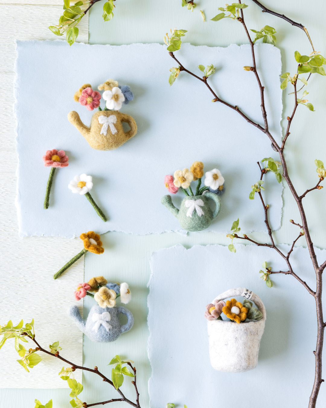 Dekorierte Szene mit Filzanhängern von Gry & Sif: Gießkannen mit bunten Blumen, einzelne Filzblumen und ein kleiner Filz-Blumentopf arrangiert auf hellblauem Hintergrund mit frischen Zweigen.