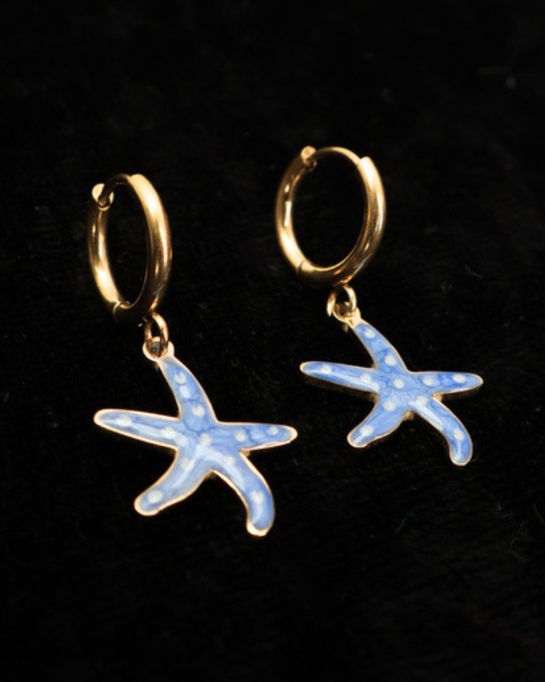 Goldene blaue Emaille-Seesterne Ohrringe von Dottilove auf schwarzer leicht glitzernder Decke.