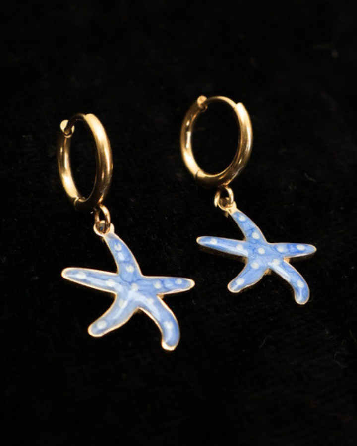 Goldene blaue Emaille-Seesterne Ohrringe von Dottilove auf schwarzer leicht glitzernder Decke.