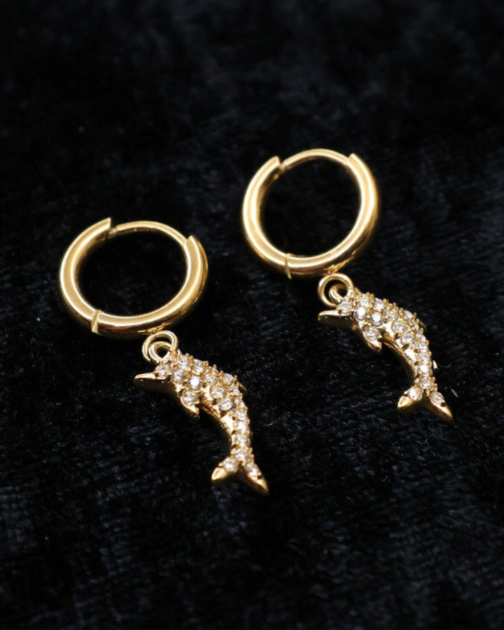 Goldene Delfin-Ohrringe von Dottilove verziert mit verschiedenen Glitzersteinen auf schwarzer leicht glitzernder Decke.