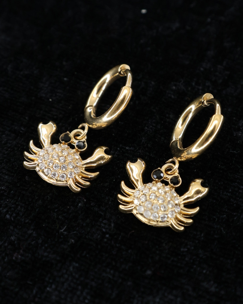 Goldene Krabbenohrringe von Dottilove verziert mit verschiedenen Glitzersteinen auf Schwarzer leicht glitzernder Decke.