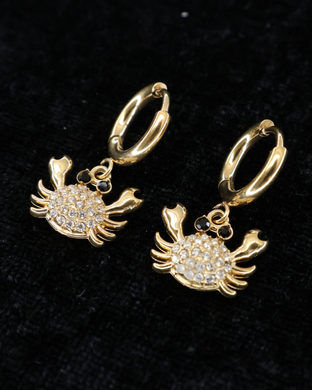 Goldene Krabbenohrringe von Dottilove verziert mit verschiedenen Glitzersteinen auf Schwarzer leicht glitzernder Decke.