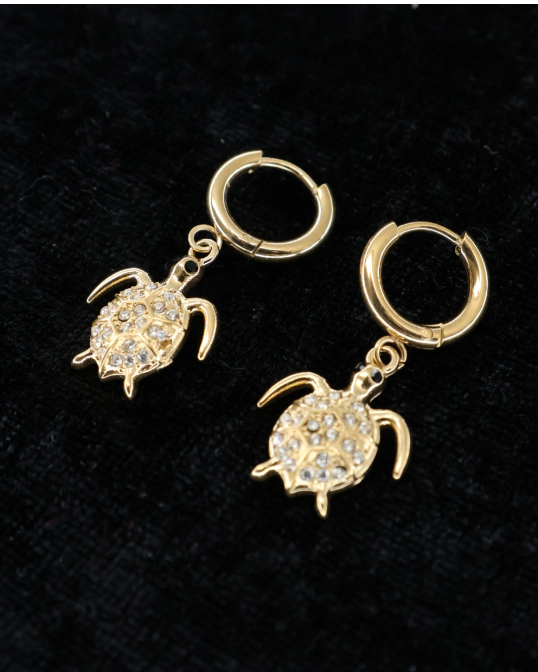 Goldene Schildkröten-Ohrringe von Dottilove verziert mit verschiedenen Glitzersteinen auf schwarzer leicht glitzernder Decke.