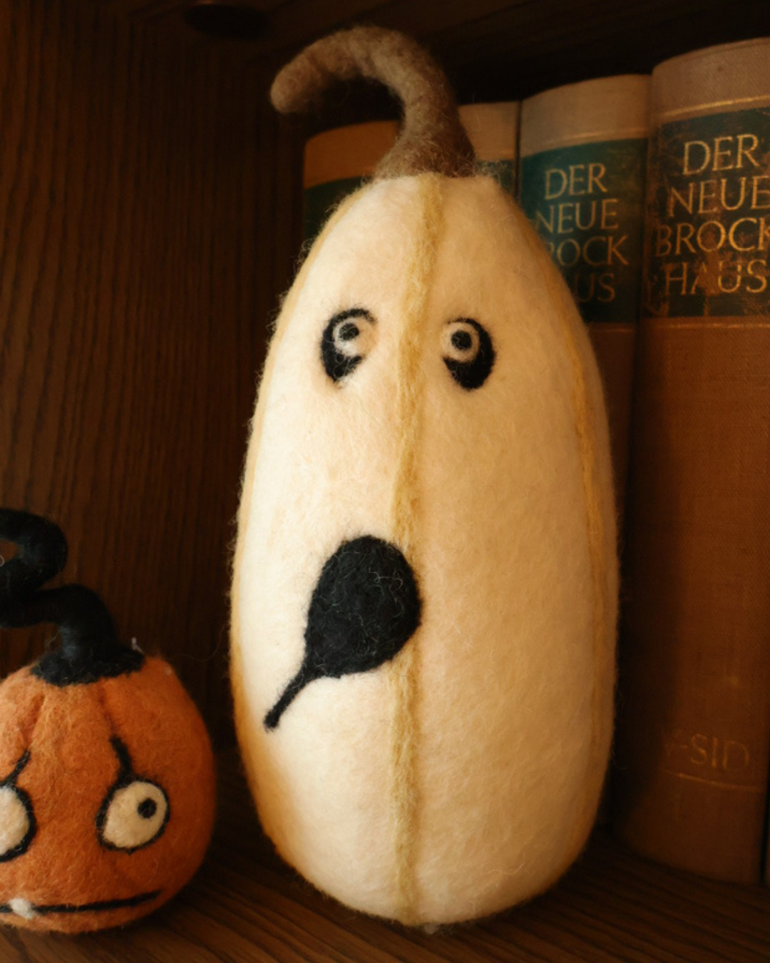Dekorierter weißer Halloween Filzkürbis von Grace & Gloria auf einem Bücherregal. Zu sehen ist ein langer weißer Kürbis mit einem schwarzen Mund schwarzen Augenringen und blauen Augen 