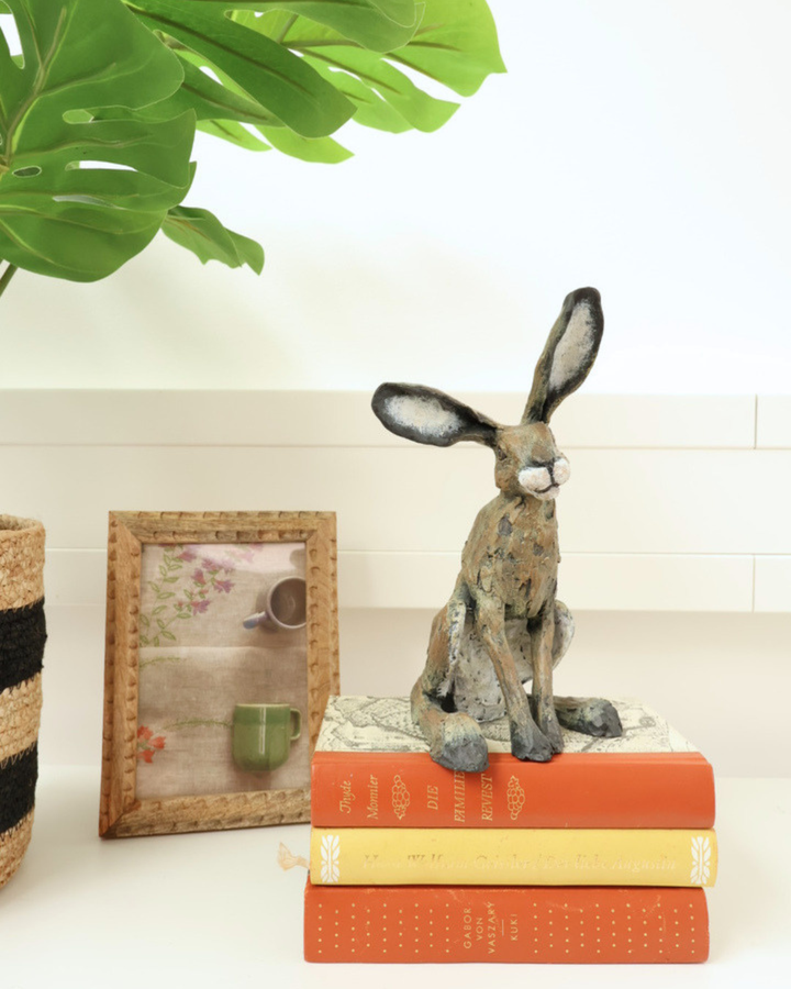 Hasen Figur "Rowan" von Wrendale Designs sitzend auf Büchern. neben dem Hasen liegt ein Bild und eine Zimmerpflanze.