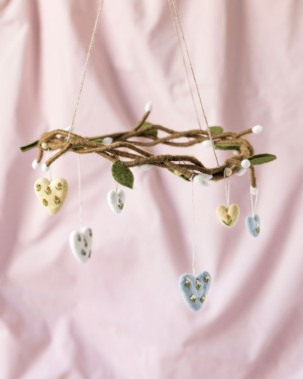 Filzanhänger aus drei bestickten Herzen von Gry & Sif, arrangiert an einem geflochtenen Zweig vor rosafarbenem Hintergrund