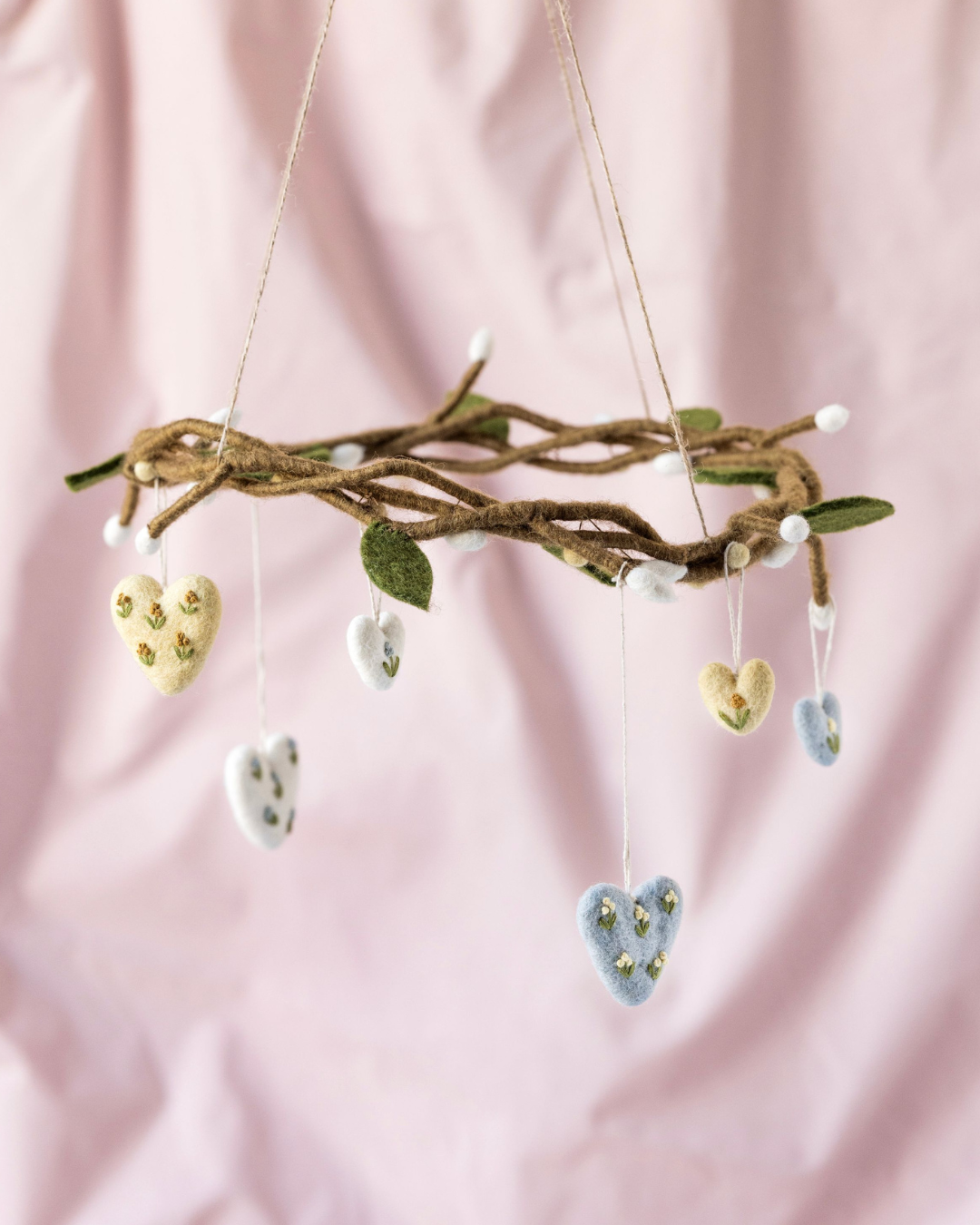 Filzanhänger aus drei bestickten Herzen von Gry & Sif, arrangiert an einem geflochtenen Zweig vor rosafarbenem Hintergrund