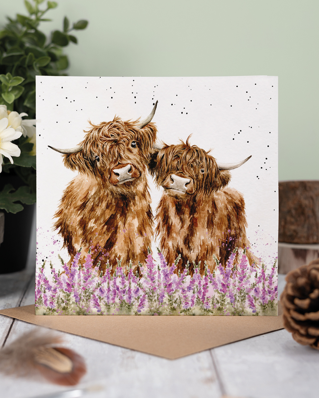 Dekoriertes Produktfoto der Hochland Kühe Grußkarte „Gentle Giants“ von Wrendale Designs, aufgestellt auf Holzuntergrund mit braunem Umschlag, im Hintergrund grüne Pflanzen, helle Blumen und natürliche Wohnaccessoires.