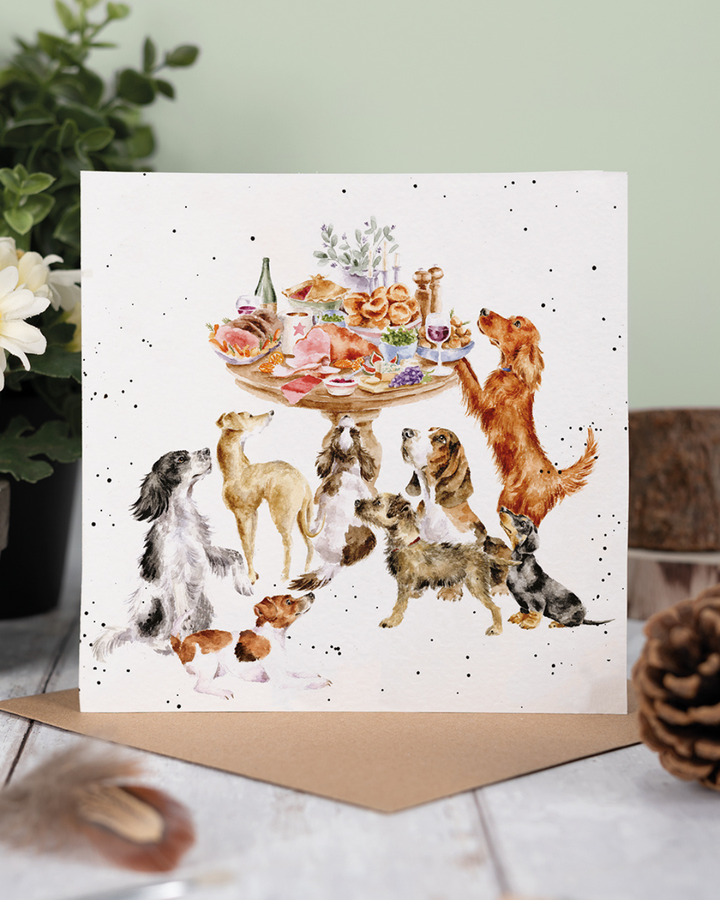 Dekoriertes Produktfoto der Hunde Grußkarte „Sunday Lunch“ von Wrendale Designs, aufgestellt vor hellgrünem Hintergrund mit Pflanzen, Holzfläche und braunem Umschlag.