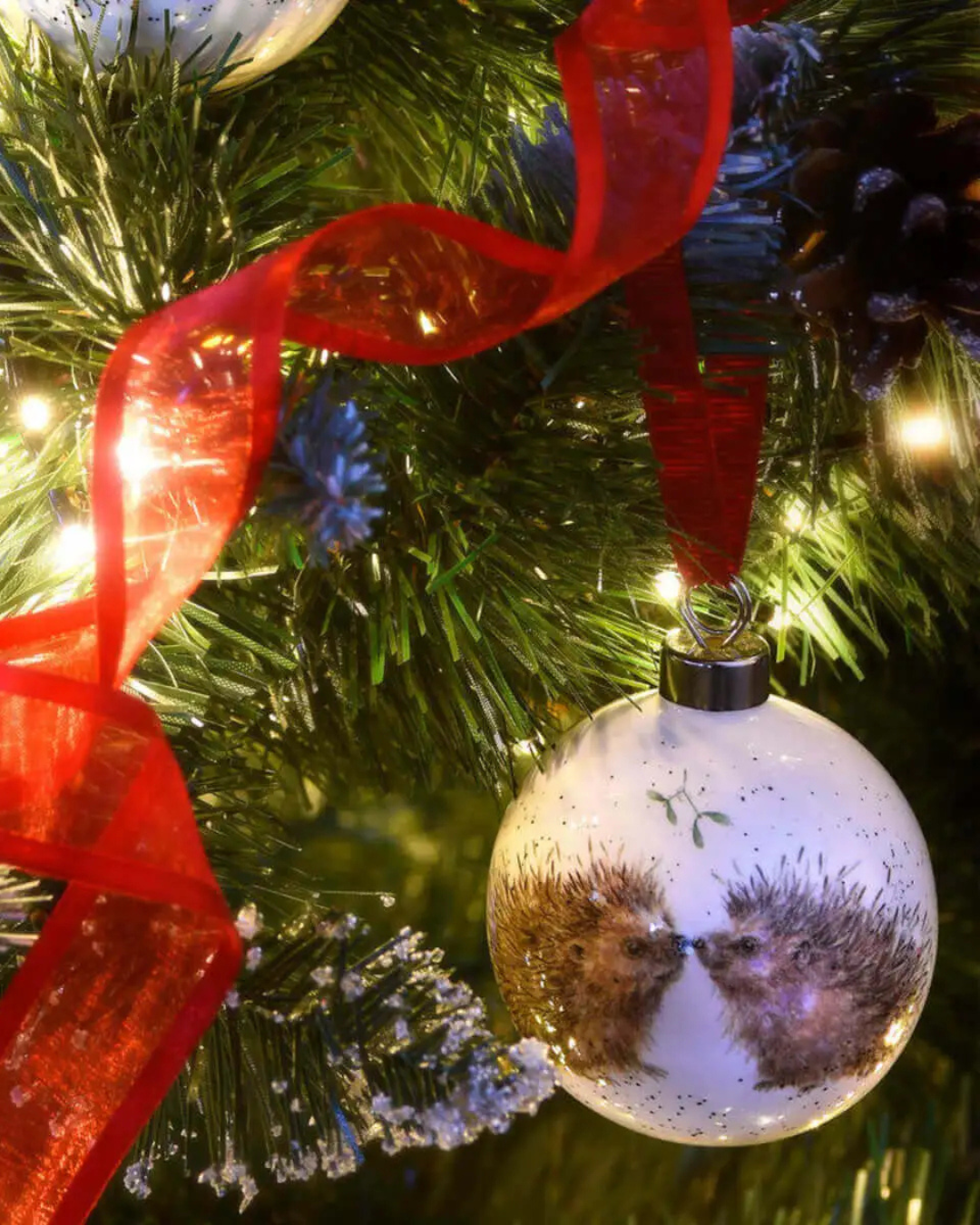Igel Porzellankugel von Wrendale Designs mit rotem Satinband am geschmückten Weihnachtsbaum.