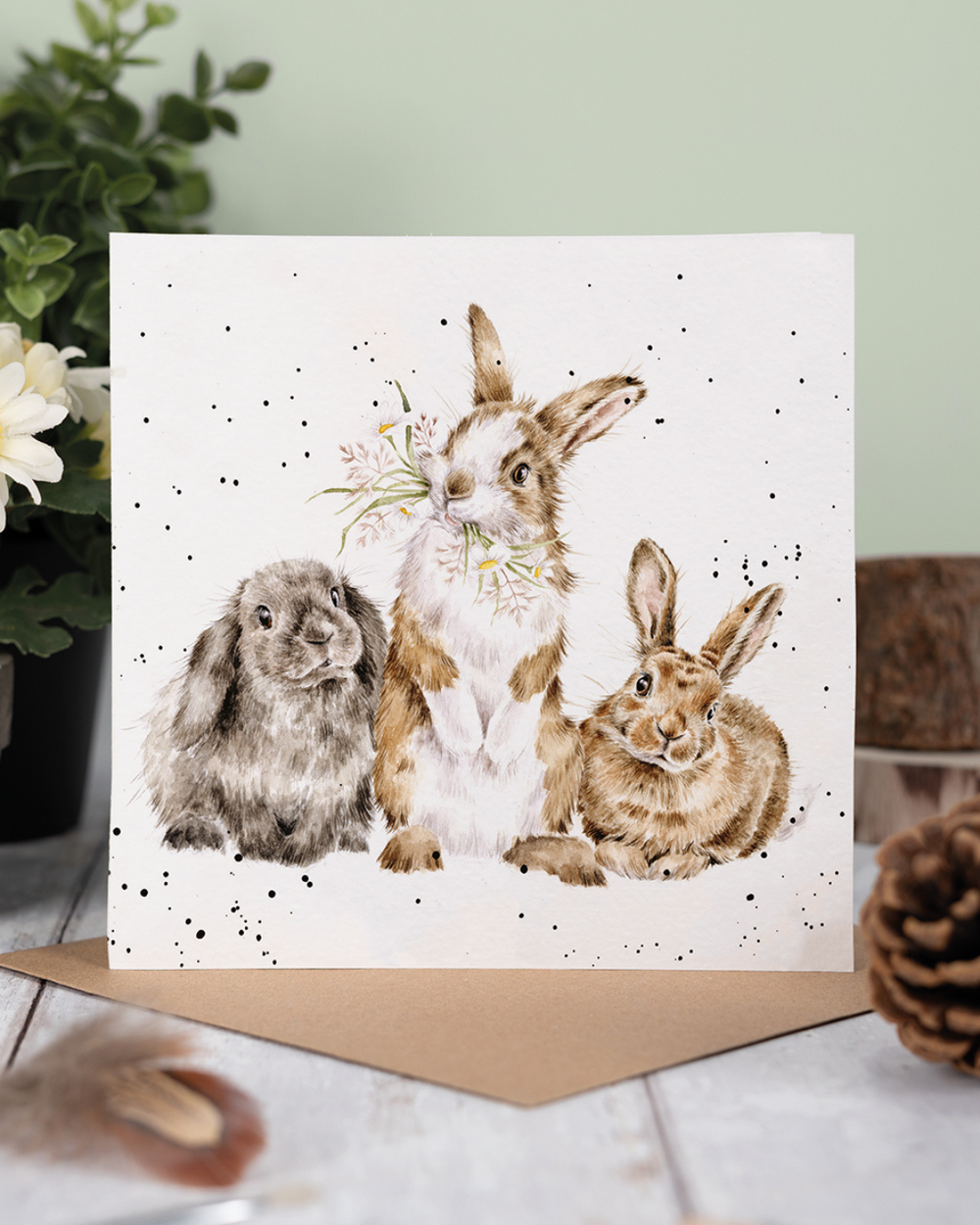 Dekoriertes Produktfoto der Kaninchen Grußkarte „All Ears“ von Wrendale Designs, stehend auf Holztisch mit braunem Umschlag, Pflanzen und natürlicher Frühlingsdekoration im Hintergrund.