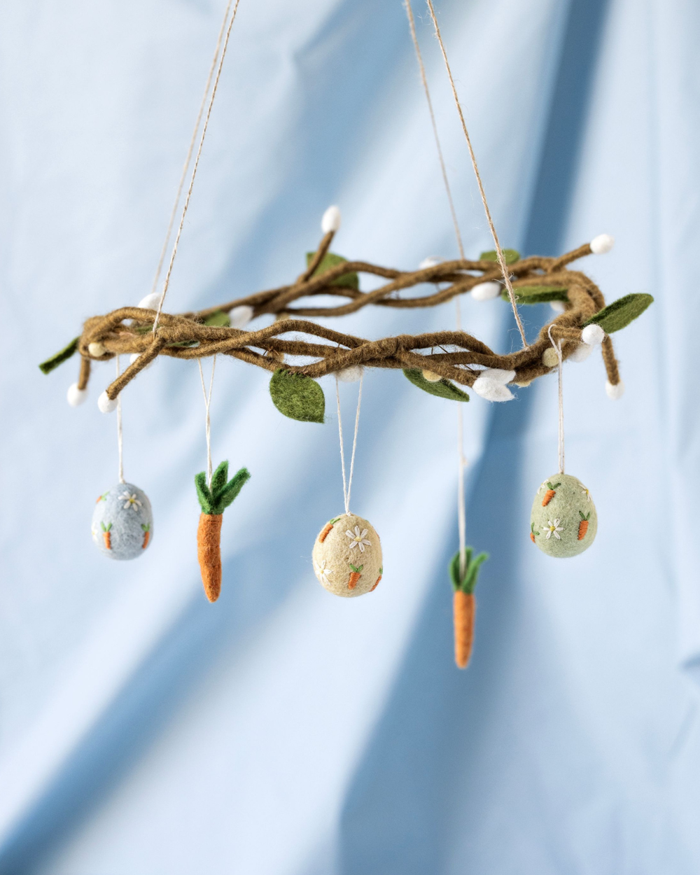 Filzkranz mit hängenden Ostereiern und Karotten-Anhängern an einer geflochtenen Filzkranz vor hellblauem Hintergrund.