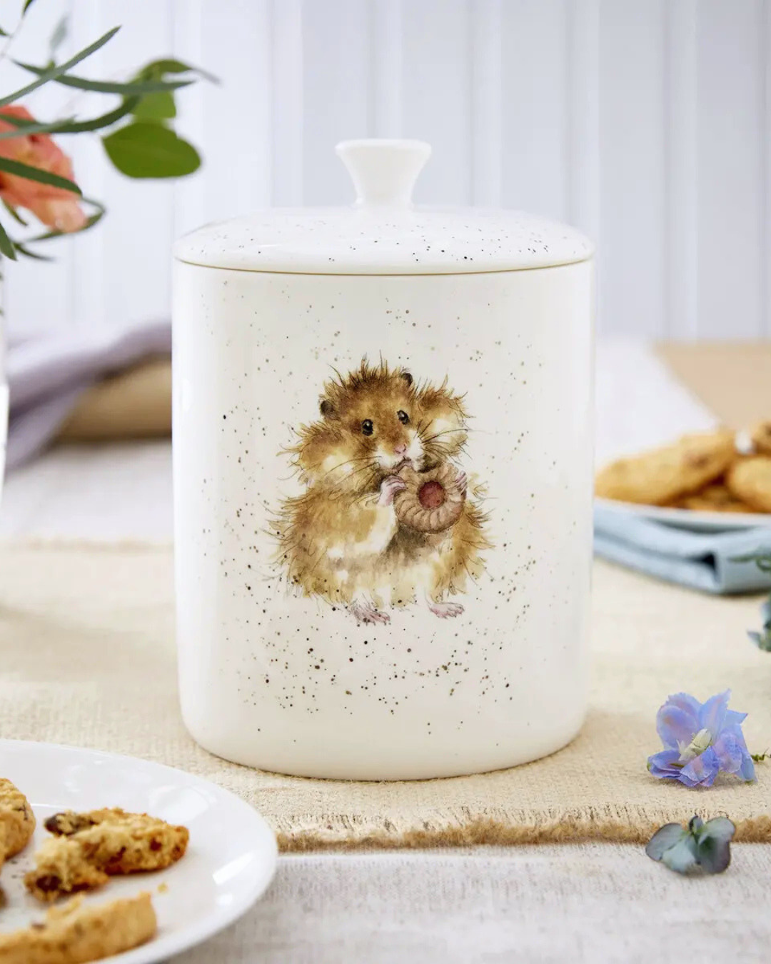 Frontansicht der Hamster Keksdose "Royal Worchester" aus Porzellan von Wrendale Designs während es auf einem bedeckten Tisch mit Keks Tellern und Blumen steht. Das Motiv zeigt einen kleinen süßen Hamster welcher an einem Keks knabbert. 