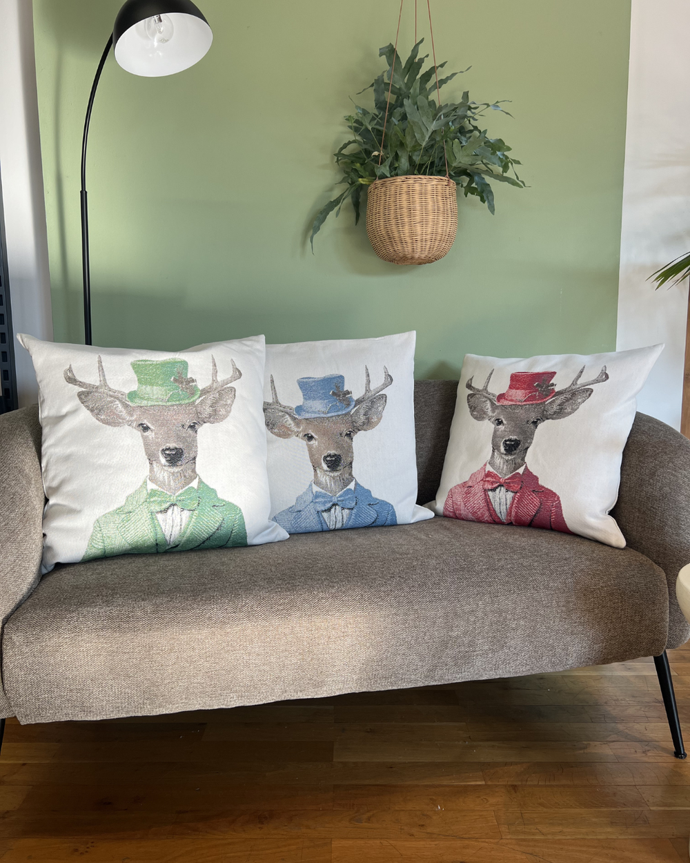 Hirschkissen "Dandy" in den drei verschiedenen Varianten/Farben liegen nebeneinander auf einer braunen Couch. Die Hirsche tragen jeweils ein helles, gestreiftes Hemd, darüber eine Weste, Anzug und  passenden Zylinder. Der linke Hirsch ist grün, der mittlere Hirsch Blau und der rechte Hirsch rot gekleidet.