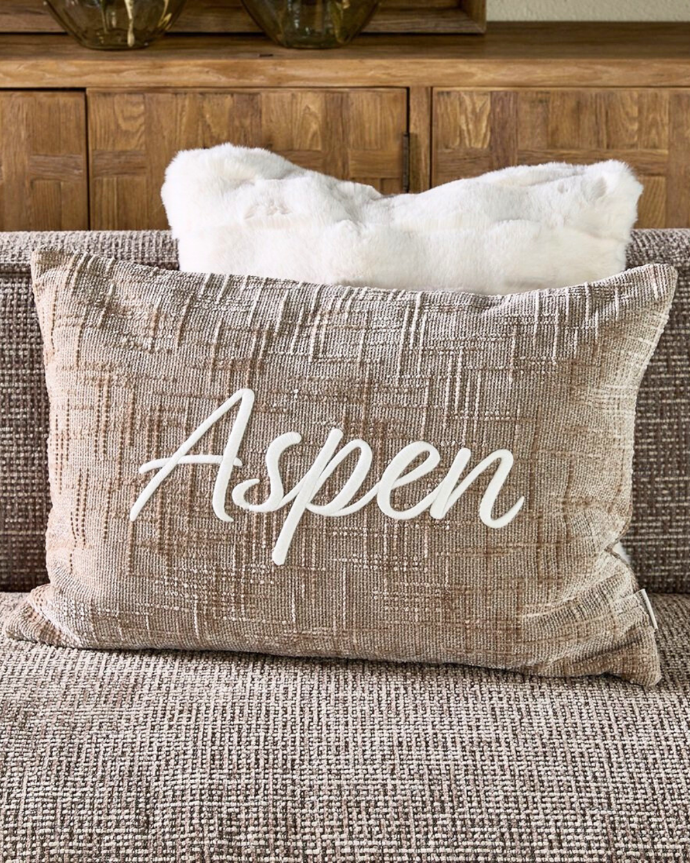 Dekorierter Aspen Kissenbezug auf braunem Sofa mit weißem Romolo Kunstfellkissen im Hintergrund.