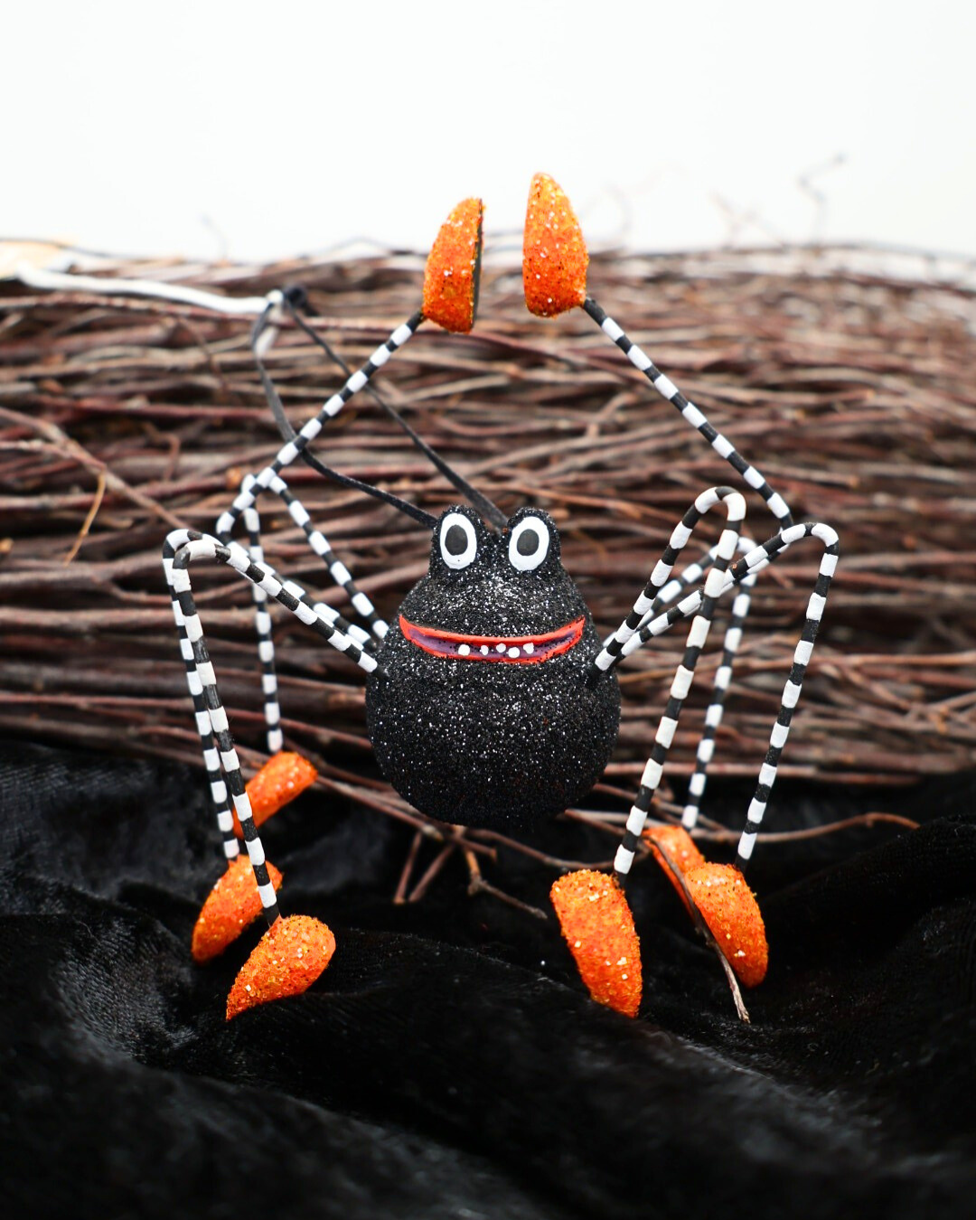 Handbemalte Halloween-Dekospinne „Webster Spider“ von Patience Brewster, mit schwarz-weiß gestreiften Beinen und orange glitzernden Schuhen, auf schwarzem Stoff vor einem Reisigbesen.