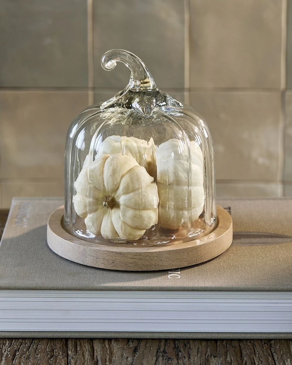 Kürbis Glasglocke "Pumpkin Cloche" von Riviera Maison mit Holzuntersetzer mit schwarzer RM Aufschrift. Hier stehen auf einem RM Dekobuch und mit drei Kürbissen gefüllt.