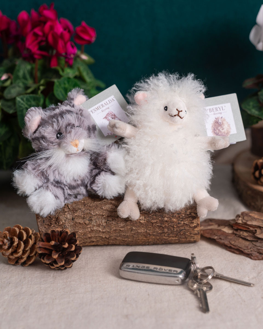 Schaf Schlüsselanhänger Beryl von Wrendale Designs zusammen mit dem Katzen Schlüsselanhänger Esmeralda auf einem Holzstück dekoriert, umgeben von Tannenzapfen und einem Autoschlüssel im Vordergrund.