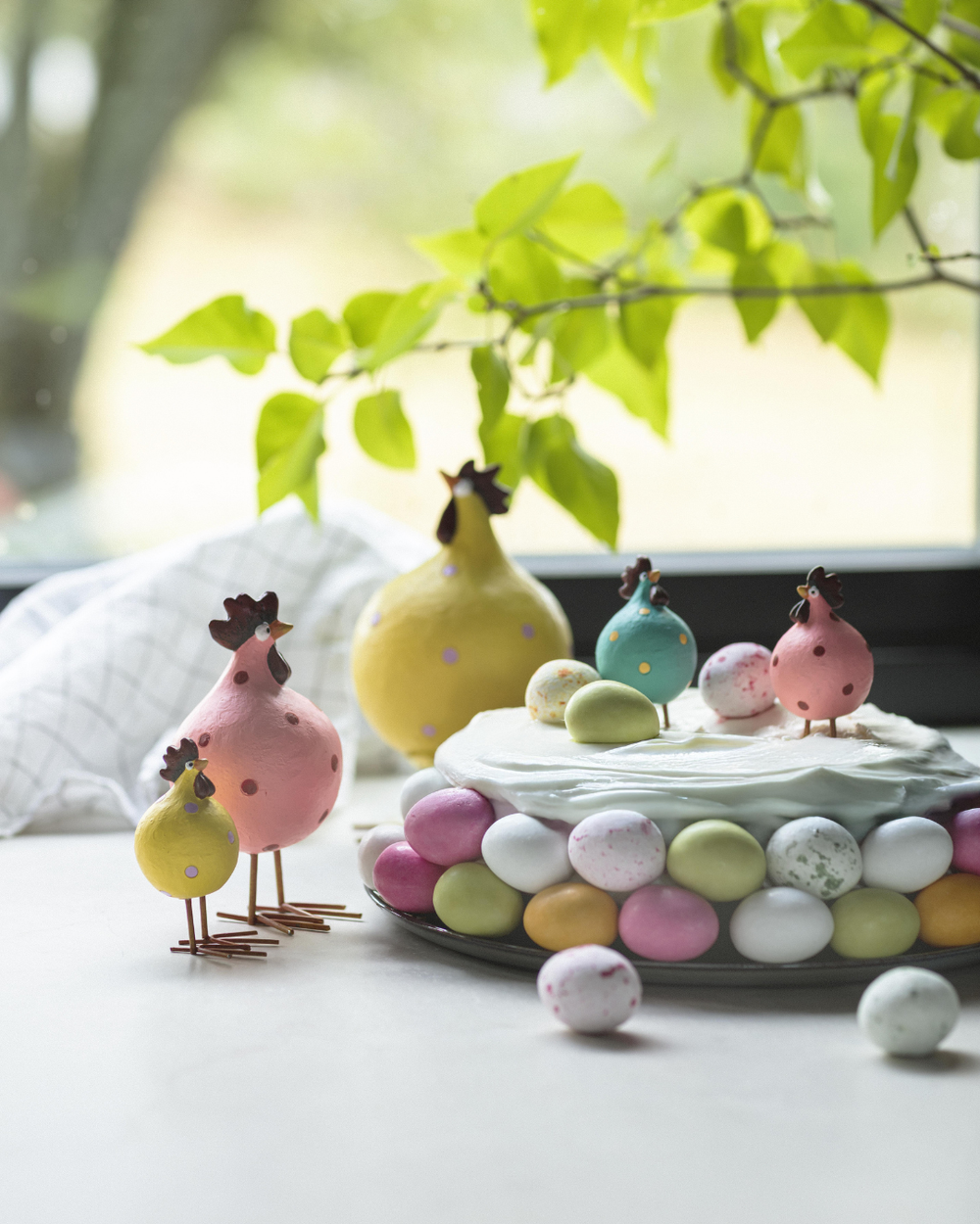 Mehrere Poppy-Hühner von Nääsgränsgården dekorativ neben Osterkuchen und bunten Eiern am Fenster arrangiert