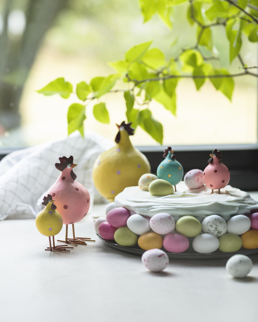 Mehrere Poppy-Hühner von Nääsgränsgården dekorativ neben Osterkuchen und bunten Eiern am Fenster arrangiert