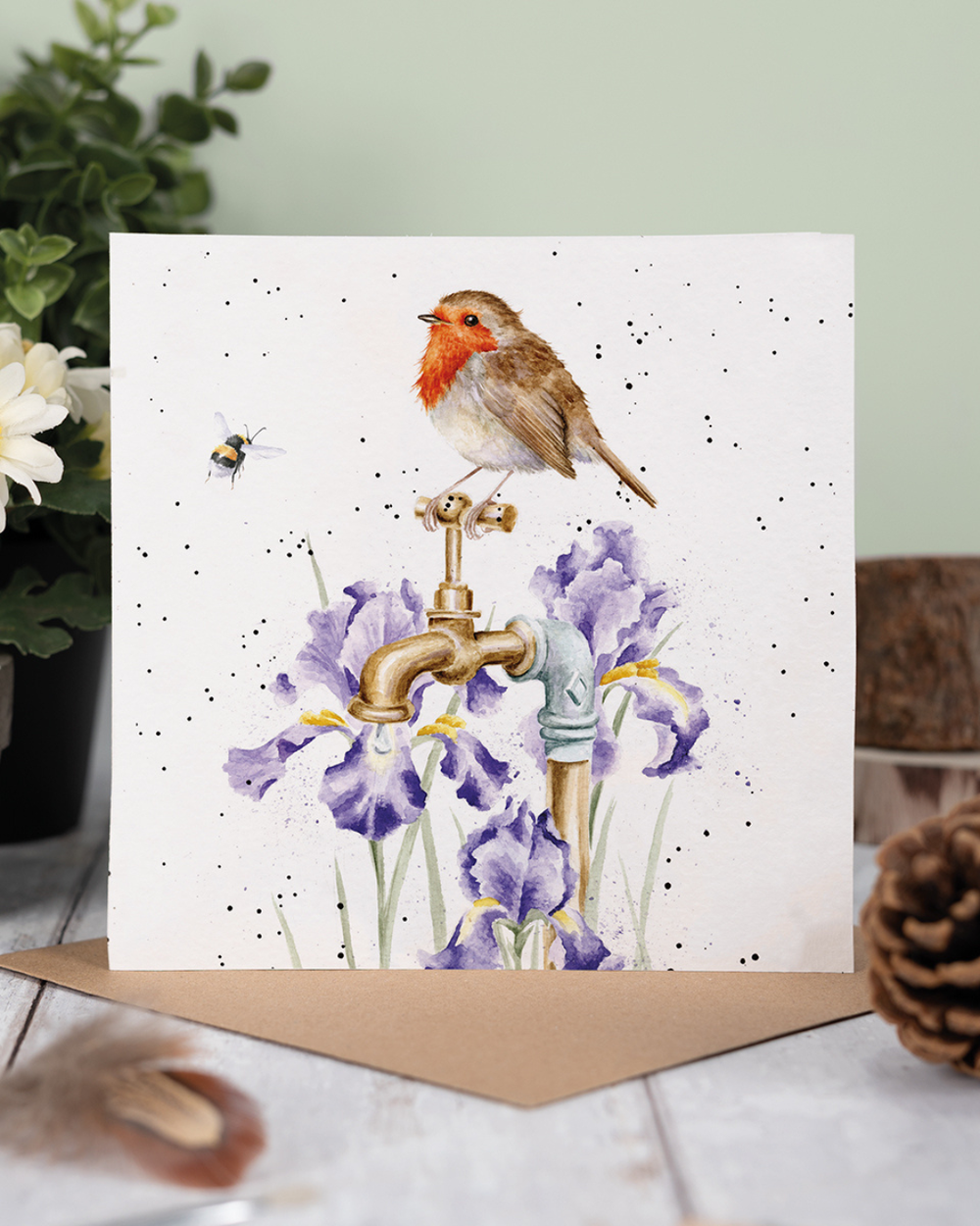 Dekoriertes Produktfoto der Rotkehlchen Grußkarte „The Gardener’s Guest“ von Wrendale Designs, mit braunem Umschlag auf Holzuntergrund, Pflanzen und natürlicher Gartendekoration im Hintergrund.