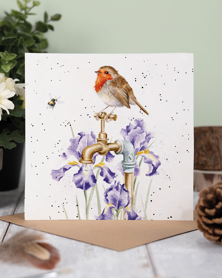 Dekoriertes Produktfoto der Rotkehlchen Grußkarte „The Gardener’s Guest“ von Wrendale Designs, mit braunem Umschlag auf Holzuntergrund, Pflanzen und natürlicher Gartendekoration im Hintergrund.