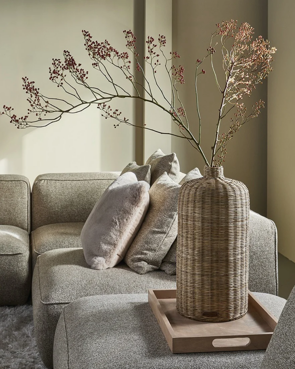 Dekorierte "Rustic Rattan Decoration Vase" von Riviera Maison in wohnlicher Atmosphäre. Die Vase steht auf einem Holztablett welches auf einem grauen Sofa liegt. 