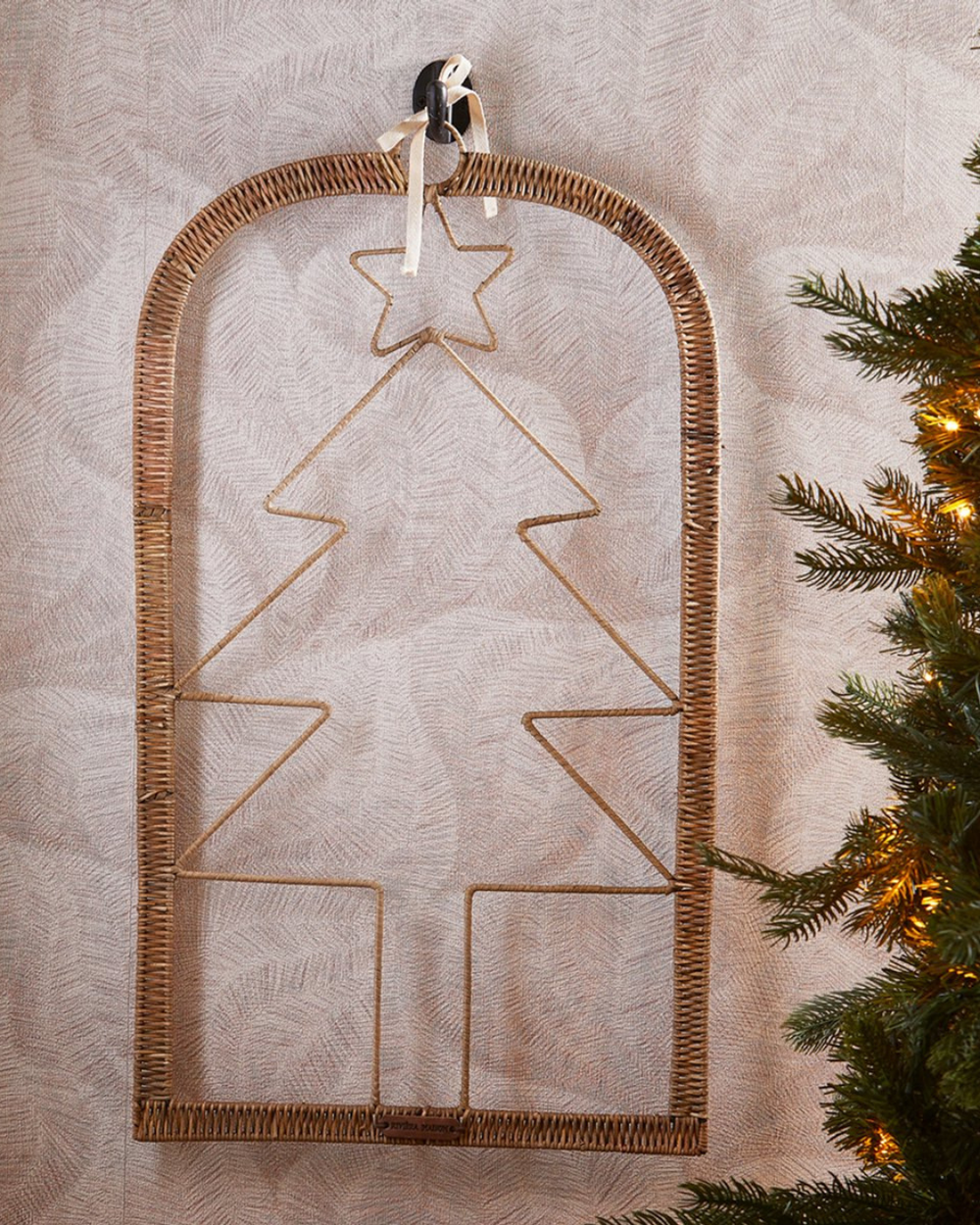 Dekorierte Wanddekoration „Rustic Rattan Xmas Tree“ von Rivièra Maison mit Tannenbaum-Form und Stern, aufgehängt neben einem Weihnachtsbaum.