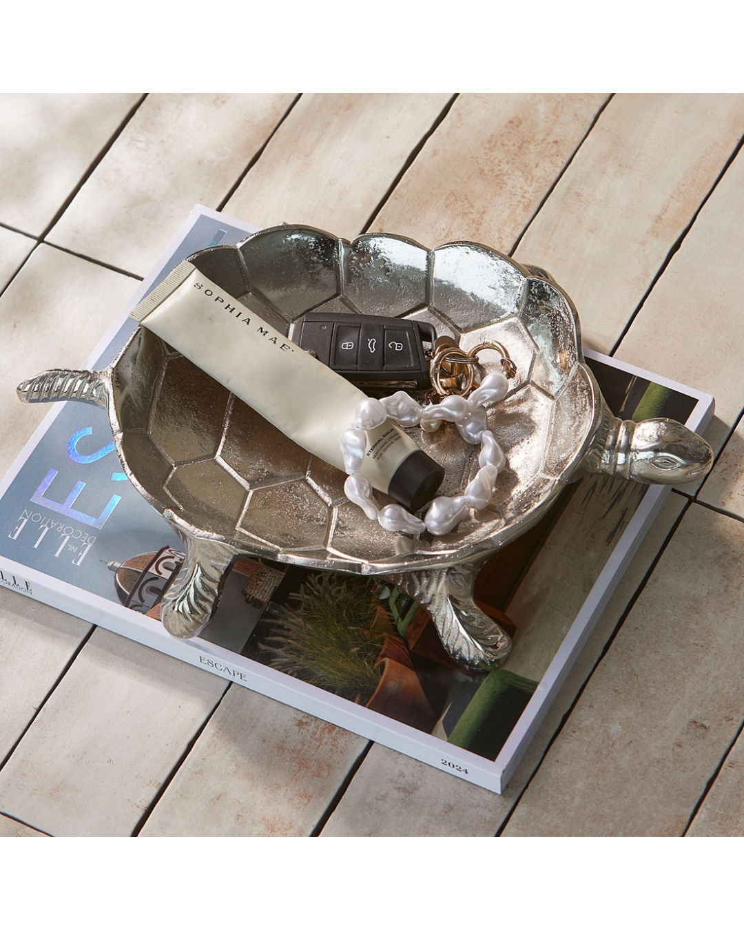 Schildkröten Dekoschale „Sea Turtle Decoration Bowl“ von Rivièra Maison dekoriert mit Schmuck und Schlüsseln, arrangiert auf einem Magazin auf hellem Holzboden.