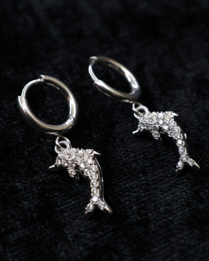Silberne Delfin-Ohrringe von Dottilove verziert mit verschiedenen Glitzersteinen auf schwarzer leicht glitzernder Decke.