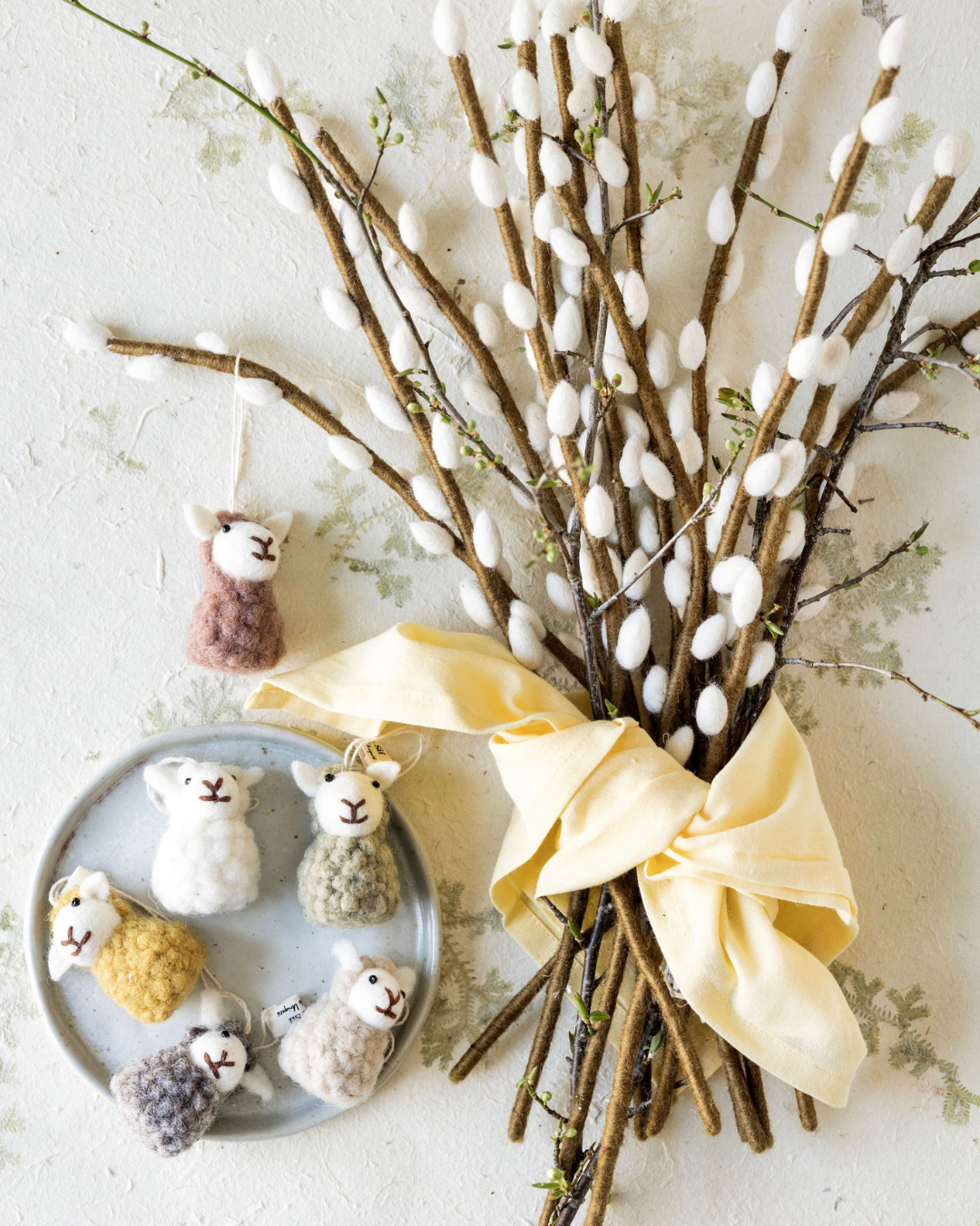 Mehrere sitzende Schaf-Filzanhänger von Gry & Sif dekorativ arrangiert an Weidenzweigen mit weißen Kätzchen, gebunden mit einem gelben Stoffband.