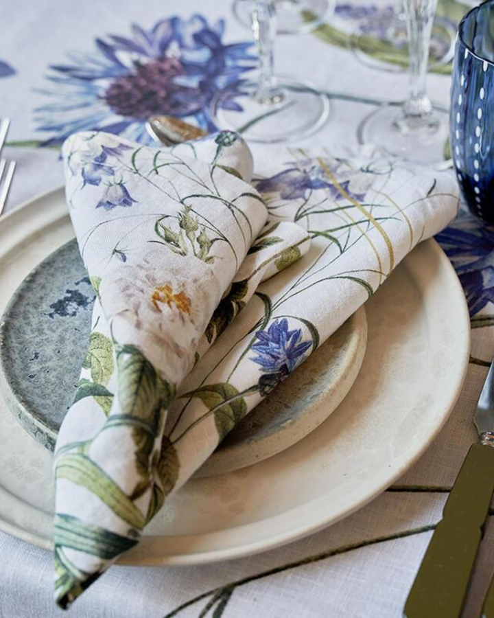 Eine gefaltete Leinenserviette „Blue Flower Garden“ von Koustrup & Co., mit einem, liegt auf einen weißen Teller mit floraler Tischdecke im Hintergrund.