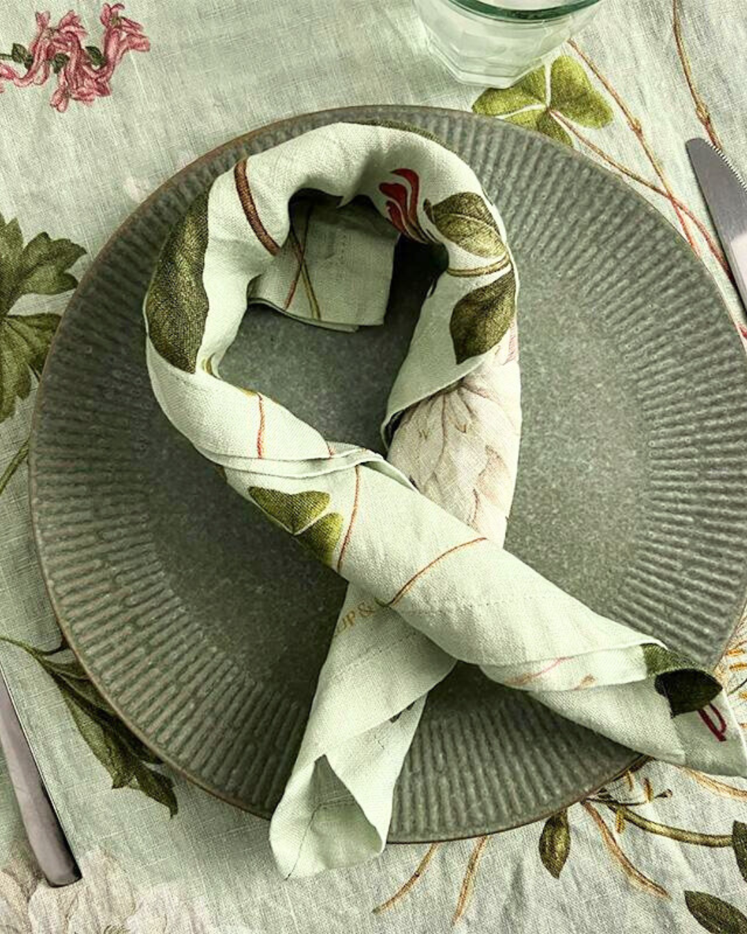 Leinenserviette "Flora Danica Atlas" von Koustrup & Co., schön gefaltet auf Tisch mit Besteck, und botanischen Motiven auf Tischdecke.