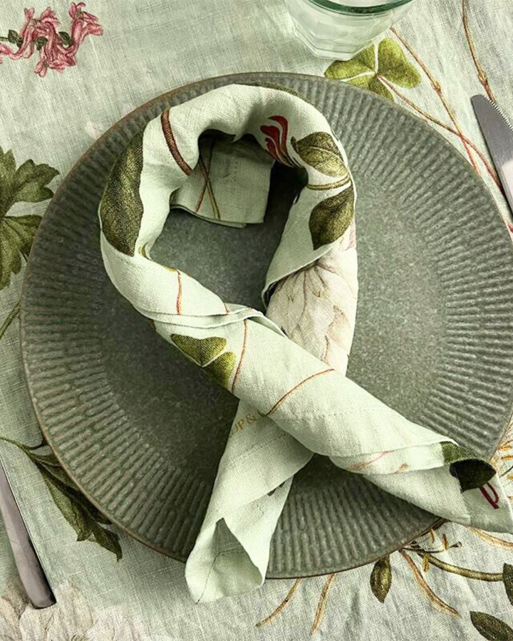 Leinenserviette "Flora Danica Atlas" von Koustrup & Co., schön gefaltet auf Tisch mit Besteck, und botanischen Motiven auf Tischdecke.