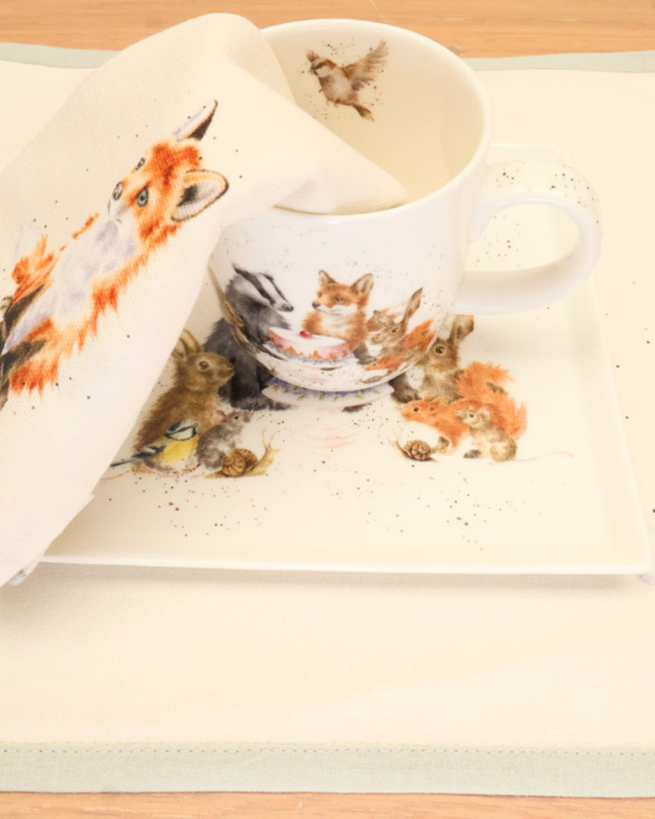 Dekorierte Serviette, Tischset, Teller und Tasse Woodland-Party von Wrendale Designs auf einem Holztisch.