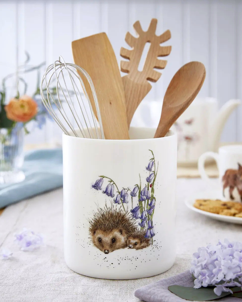 Igel Utensilien Aufbewahrung "Royal Worchester" aus Porzellan von Wrendale Designs auf einem gedeckten Tisch während es mit Kochlöffeln etc. gefüllt ist. Das Produkt ist größtenteils weiß. Das Motiv zeigt einen großen und einen kleineren Igel welche neben 2 Pflanzen mit einander kuscheln.