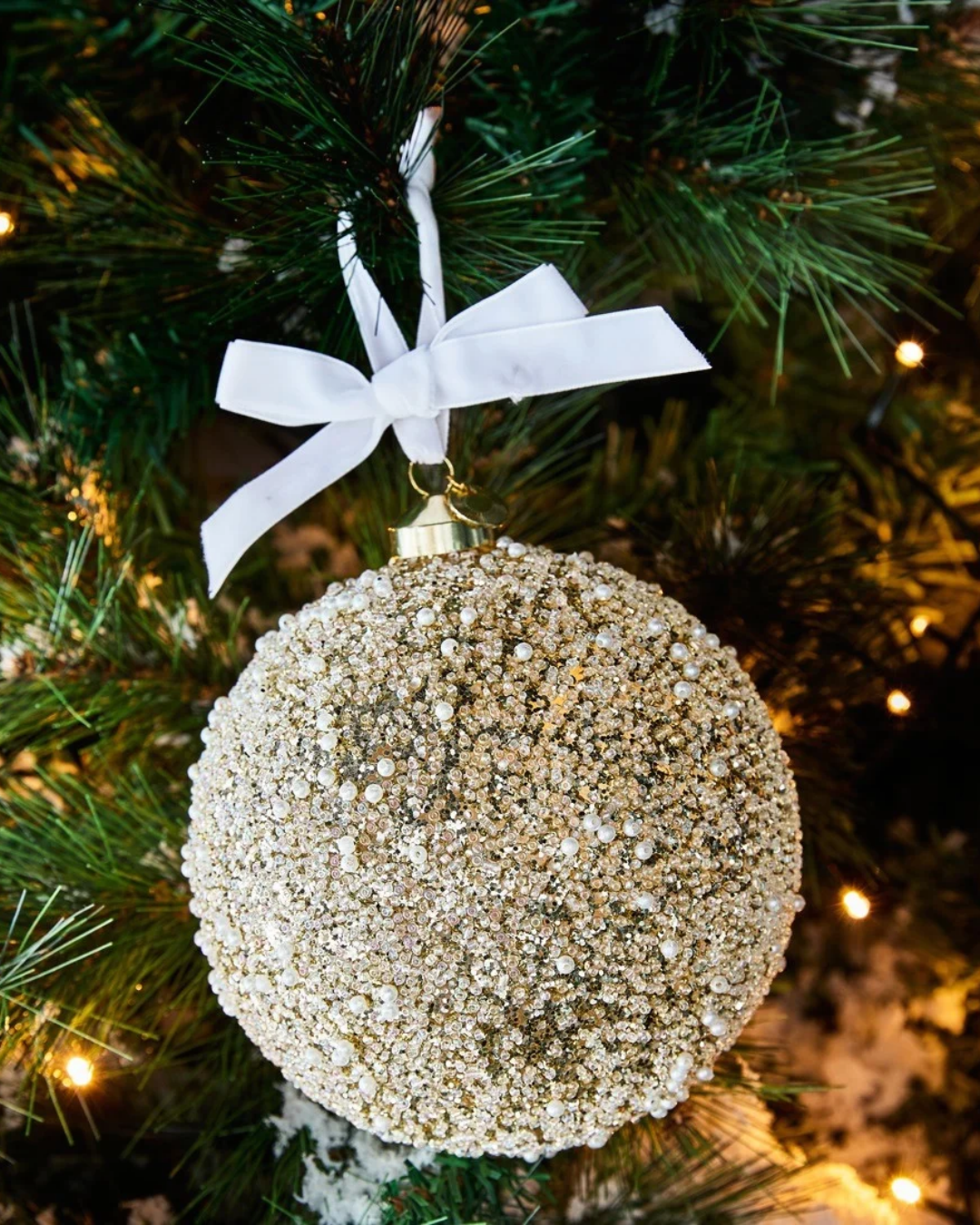 Dekorierte Weihnachtsbaumkugel "Sparkle Shine Ornament" von Riviera Maison. Das glitzernde goldene Ornament hängt mit einer weißen Schleife an einem leuchtenden Tannenbaum. 