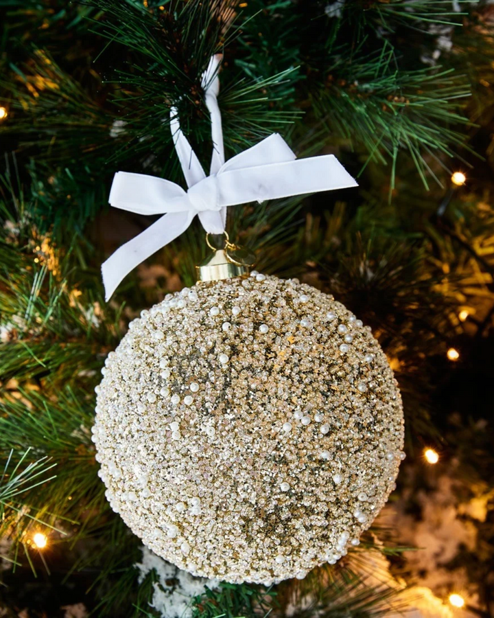 Dekorierte Weihnachtsbaumkugel "Sparkle Shine Ornament" von Riviera Maison. Das glitzernde goldene Ornament hängt mit einer weißen Schleife an einem leuchtenden Tannenbaum. 