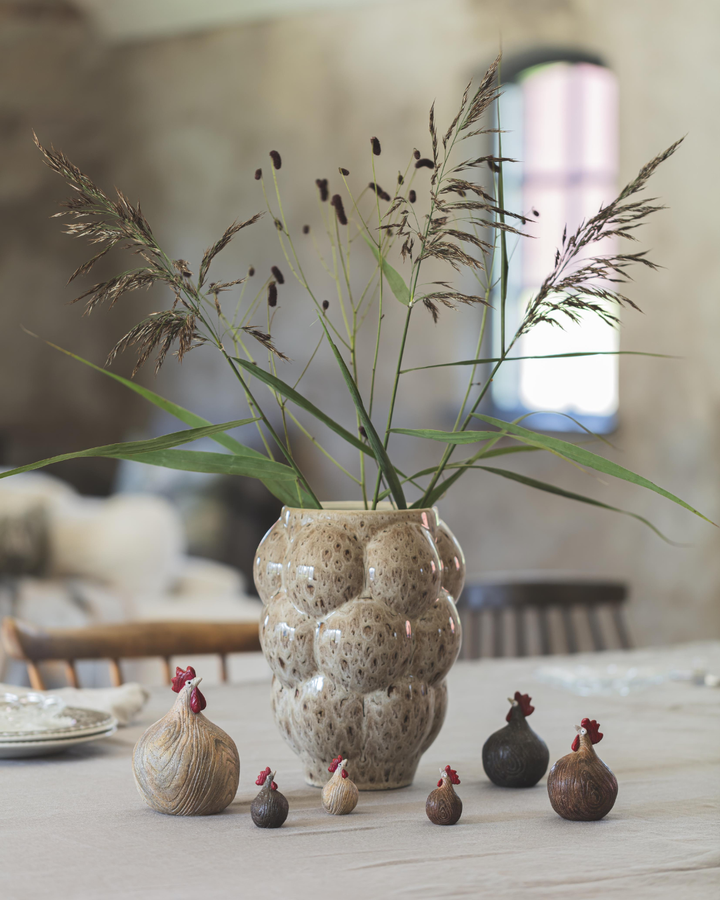 Mehrere Hühner „Woodie“ von Nääsgränsgården dekorativ auf einem Tisch vor Vase mit Gräsern arrangiert