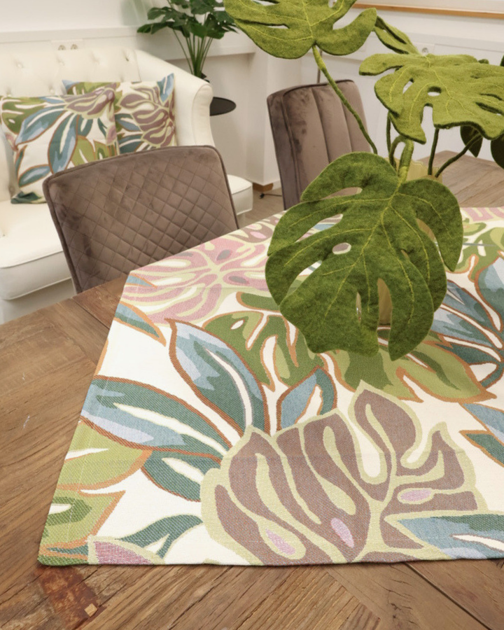 Zimmerpflanzen Mitteldecke Selva von Sander Table + Home. Die mehrfarbigen Monsterblätter sind sich gegenseitig überlappend dargestellt. Blaue, grüne und roséfarbene Blätter mit sich farblich absetzender Blattumrandung sind in den Stoff der Mitteldecke gewoben. Im Hintergrund sind die Zimmerpflanzen Kissen Selva auf einer weißen Couch liegend zu sehen.