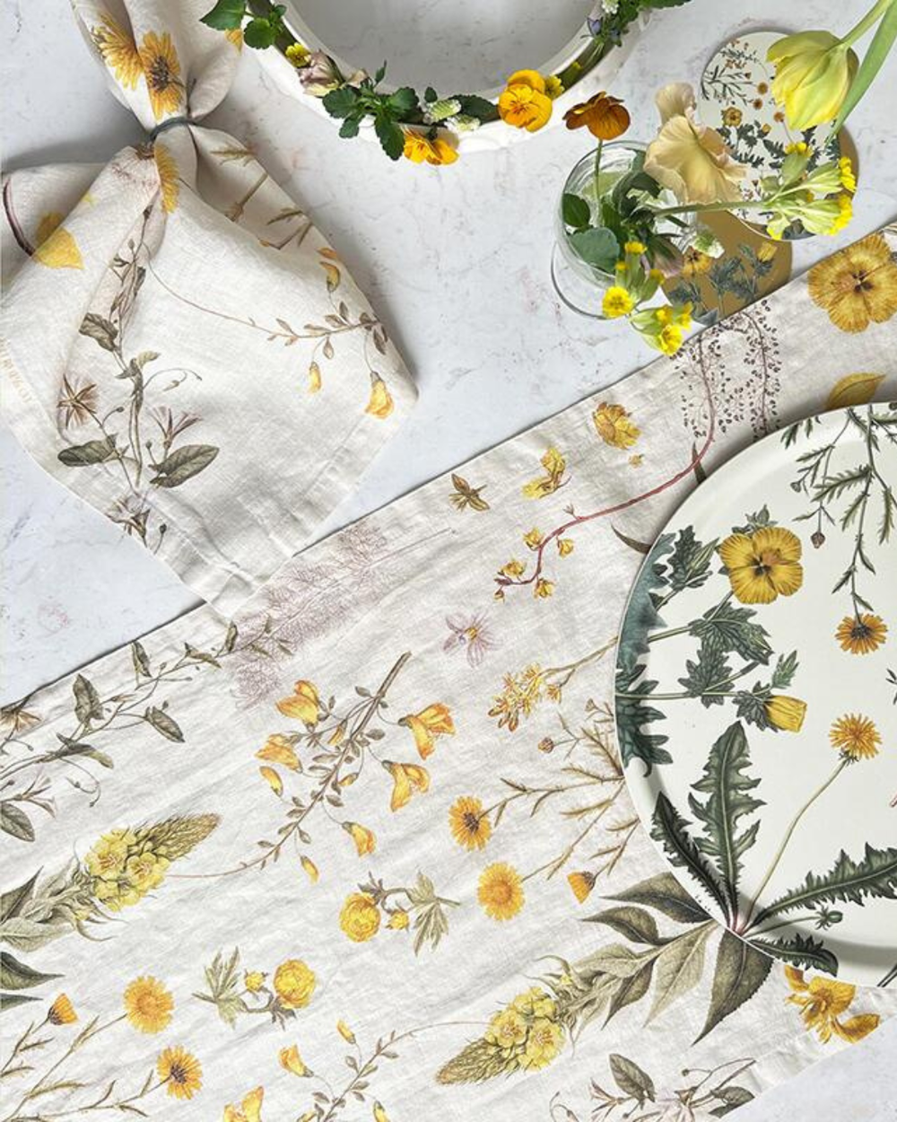 Tischläufer "Flora Danica Atlas – gelb" mit Blumenmotiv gedeckt auf weißen Tisch. Auf dem Tisch stehen: Stoffserviette, Teller, Kranz und Blumenvasen alle in einem ähnlichen Design.