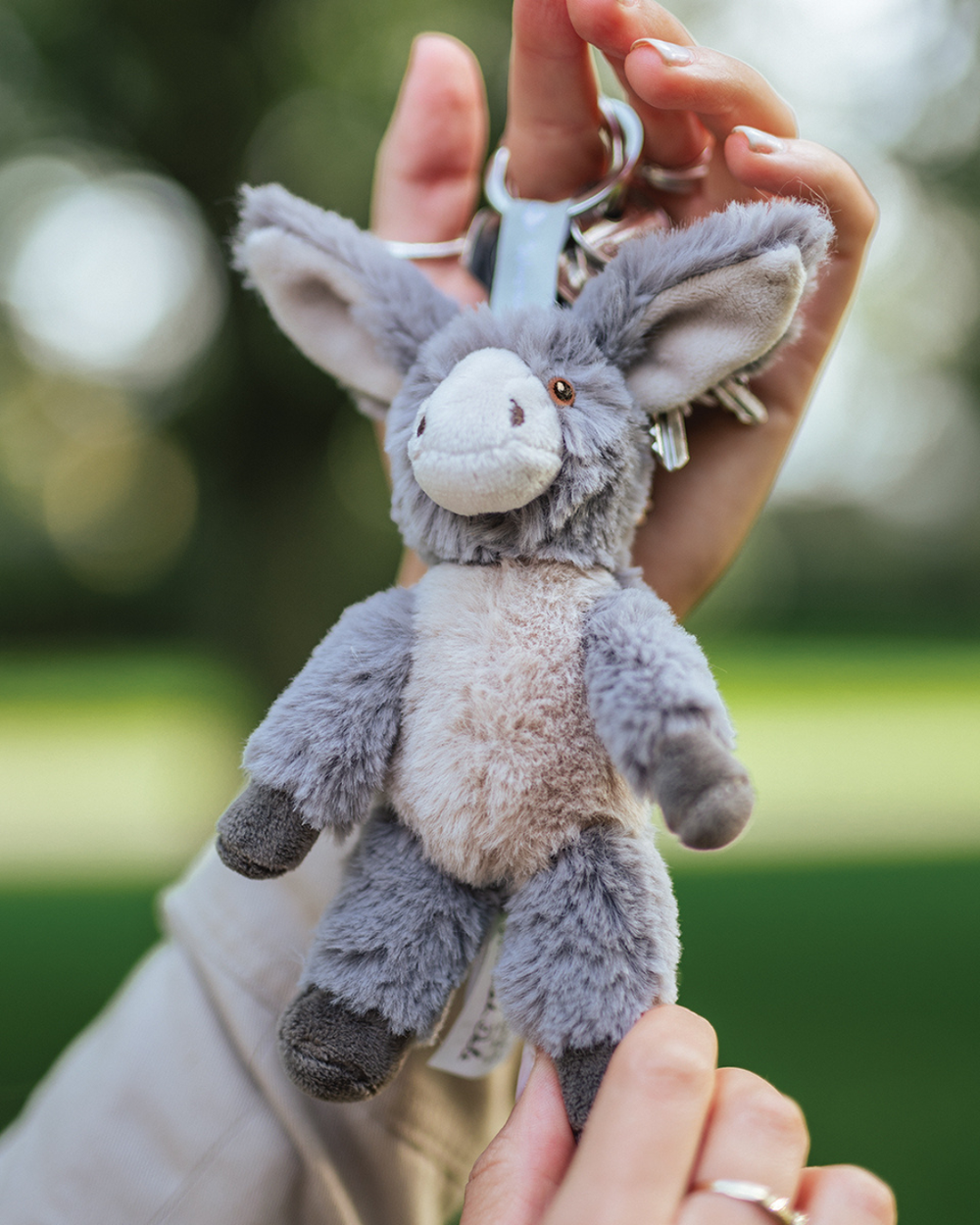 Grauer Esel Schlüsselanhänger Jack von Wrendale Designs, an einem Schlüsselring in einer Hand gehalten, mit großen Ohren und weichem Fell vor unscharfem grünen Hintergrund im Freien.