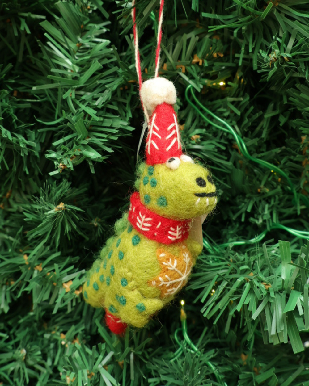 Dekorierter grüner Dino Weihnachts-Anhänger von Grace und Gloria. hängt an einem Tannenbaum. Der Dino ist hellgrün mit dunklen Punkten trägt eine Weihnachtsmütze ein Schal und hat viele Weihnachtliche Details.