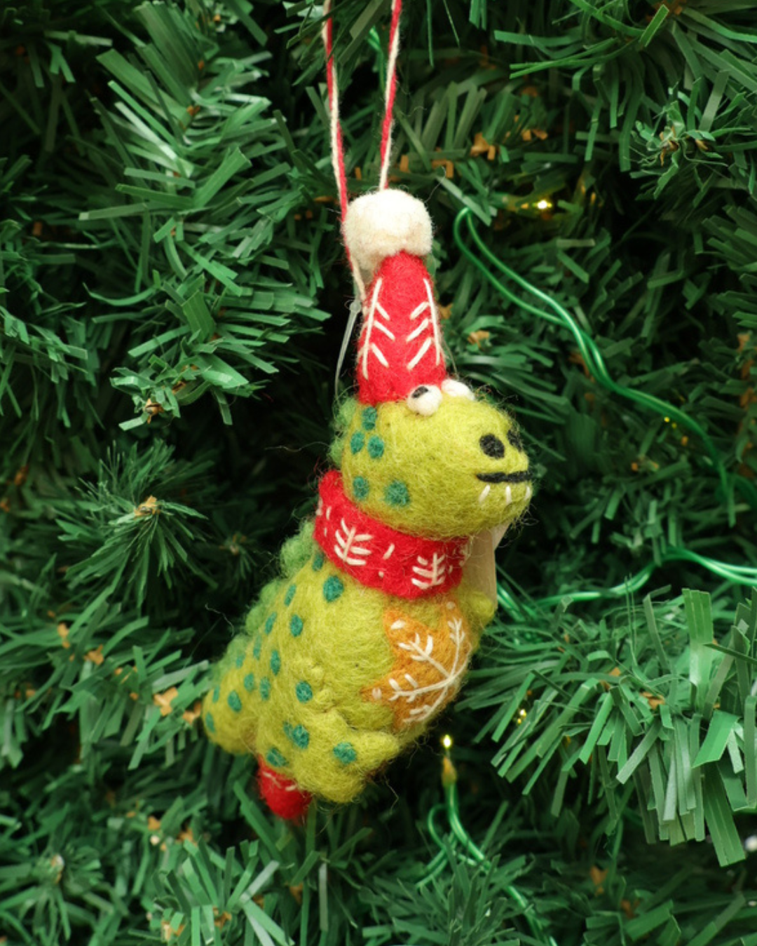 Dekorierter grüner Dino Weihnachts-Anhänger von Grace und Gloria. hängt an einem Tannenbaum. Der Dino ist hellgrün mit dunklen Punkten trägt eine Weihnachtsmütze ein Schal und hat viele Weihnachtliche Details.