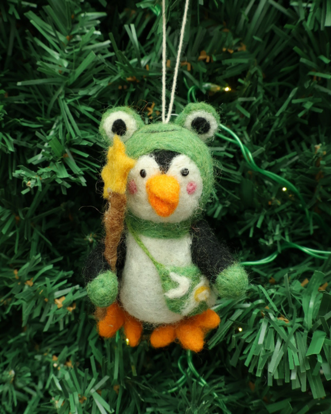 Dekorierter Pinguin Weihnachts-Filzanhänger im Froschkostüm hängt an einem Tannenbaum. Der Pinguin hat rote Wangen und viele grüne Details.