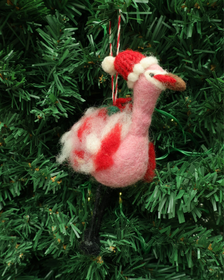Dekorierter Flamingo Weihnachts-Filzanhänger "Jutta" hängt an einem Tannenbaum. Der Flamingo ist pink, trägt eine Weihnachtsmütze und hat viele Weihnachts-Details.