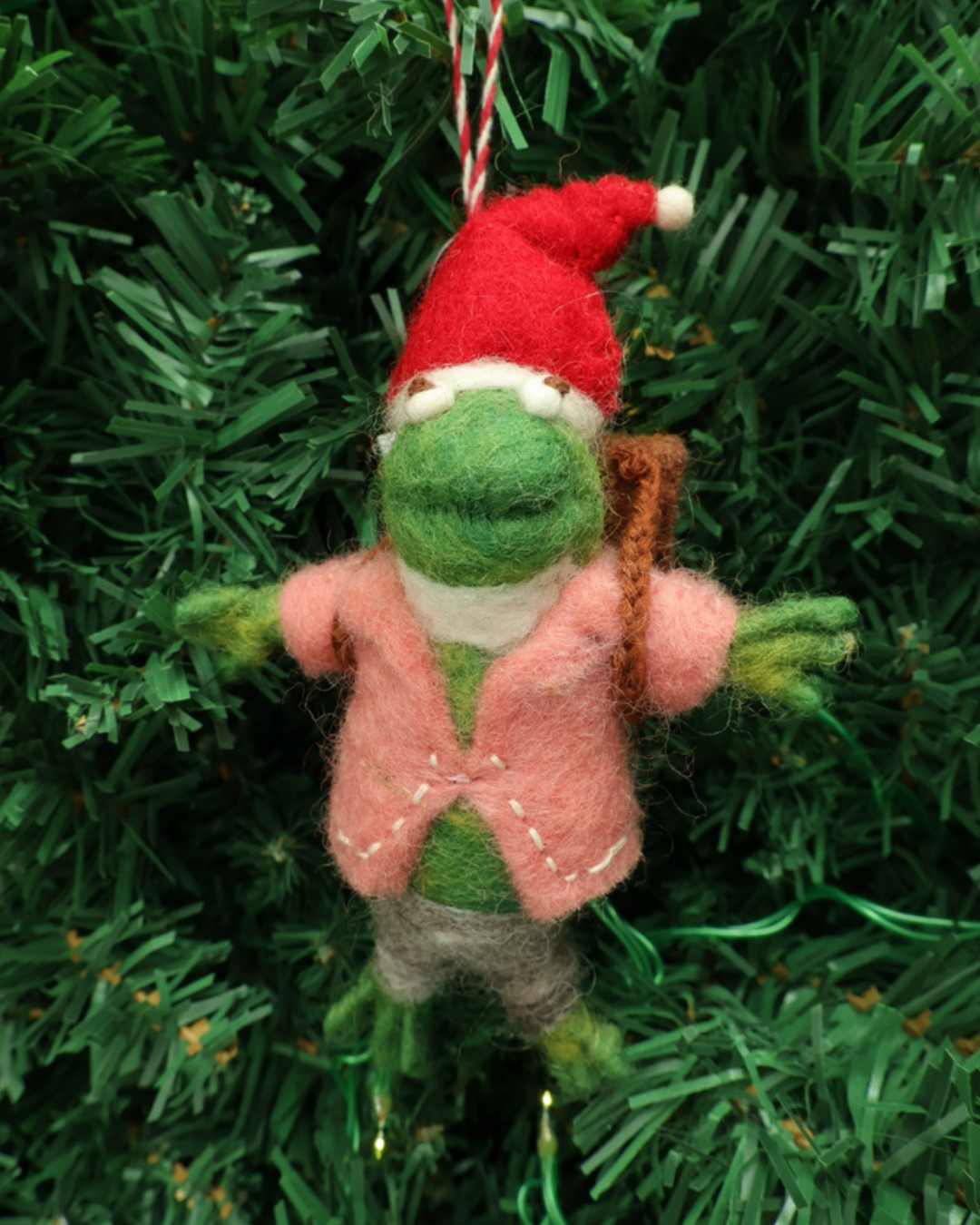 Dekorierter Frosch Weihnachts-Filzanhänger "Pit" von Grace und Gloria hängt an einem Tannenbaum. Der Frosch trägt ein pinkes Oberteil, eine graue Hose und einen roten Weihnachtshuts.