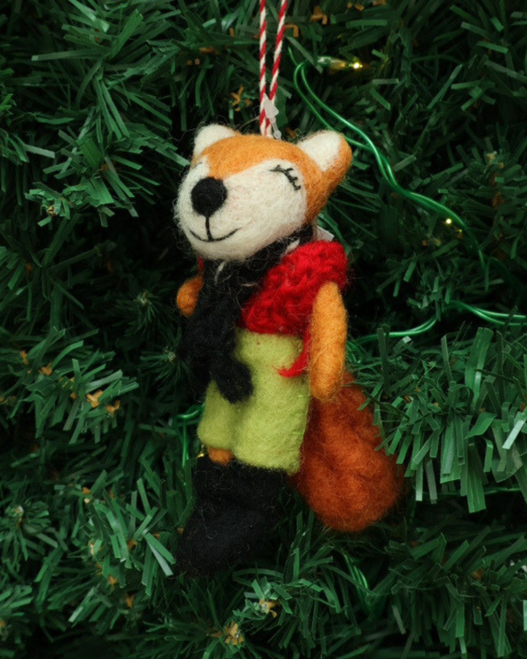 Dekorierter Weihnachts-Fuchs-Filzanhänger "Nils" von Grace & Gloria hängend an einem Tannenbaum. Es ist ein süßer Fuchs mit einer grünen Hose und einem roten Kleid.